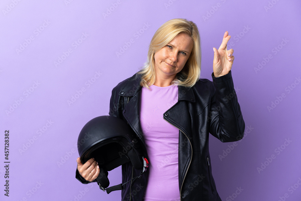 Wall mural Middle age woman holding a motorcycle helmet isolated on purple background with fingers crossing and wishing the best