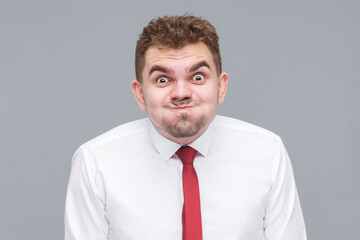funny portrait of young man in white shirt and tie standing blowing his cheeks and looking at camera with funny face and big eyes. indoor isolated on gray background.
