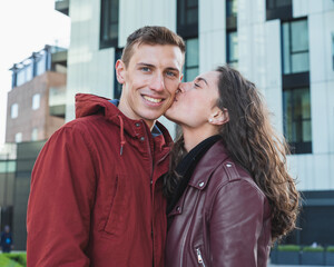 Young couple in tender attitude, girl kissing boy on the cheek, background modern buildings, love concept of the generation millenials