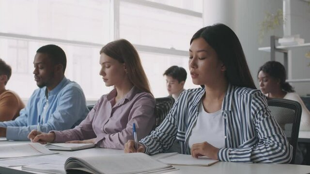 Lecture at college. Group of international students writing notes during lesson, sitting together at light classroom