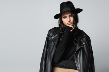 young woman in fedora hat, black turtleneck and trendy leather jacket posing isolated on grey