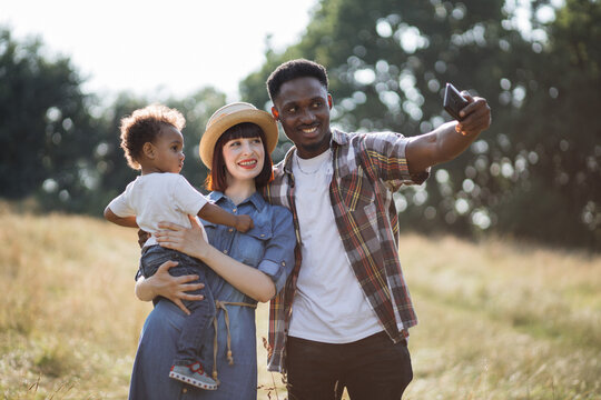 Positive multiracial family of three spending summer time on nature and taking selfie for saving memories. Young parents with cute son using modern smartphone on fresh air.