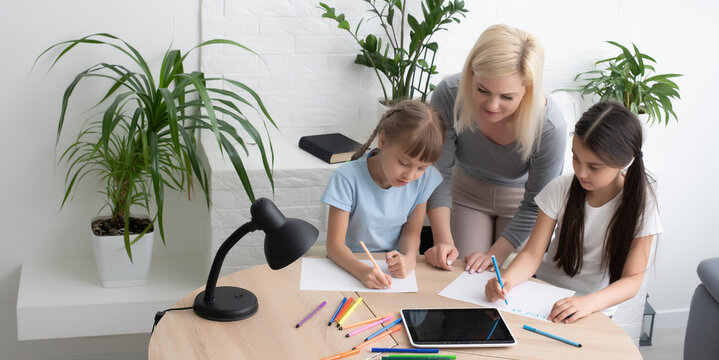 Woman tutor or foster parent mum helping cute caucasian school child girl doing homework sitting at table. Diverse nanny and kid learning writing in notebook studying at home - Powered by Adobe