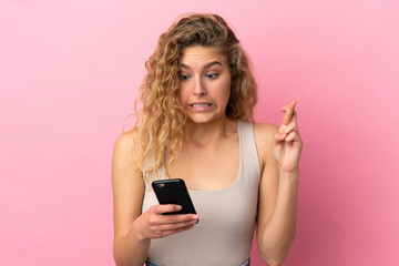 Young blonde woman isolated on pink background using mobile phone with fingers crossing