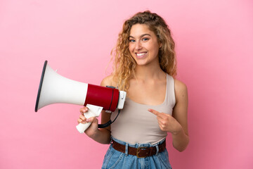 Young blonde woman isolated on pink background holding a megaphone and pointing side