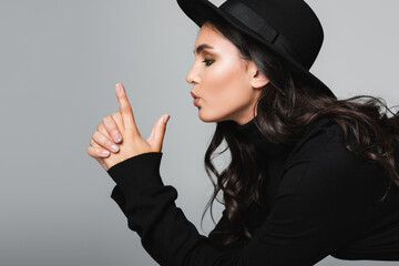 side view of brunette model in fedora hat pouting lips and showing finger gun gesture isolated on grey