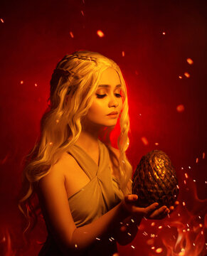 Fantasy blond woman queen holds in her hands a dragon egg. Girl elf princess looks with hopefully love. Background red flaming fire, smoke and sparks. Studio creative photography mixed red light.