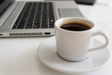 The concept of a workplace with coffee in a neat white mug, a laptop and a notebook with a pen on a modern white office desk. Easy to use. Minimalism. Side view.