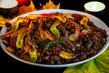 Dish for decorating table Halloween. In white plate marmalade worms in ground made of crumbled chocolate cookies. Halloween food, decor. Dirt cups with gummy worms, Halloween dessert for kids.
