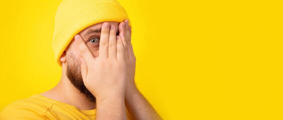 Handsome man hides face with hands, looking through palms, feels shy over yellow background,...