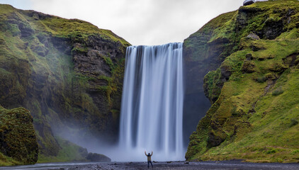 a lone male traveler experiencing the breathtaking and powerful Skogafoss waterfall in Iceland. scale of how huge the waterfall compared to the size of a man.