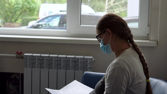 Young Pregnant Woman In Medicine Protective Mask Reading On Couch Waiting For Appointment Medical Control On Hospital. Visit Gynecologist Doctor At Clinic. Examine Expectant Belly Healthcare Check Up