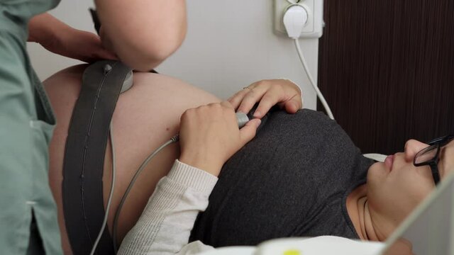 Pregnant Woman On Hospital Couch During Medical Control Cardiotocography. Gynecologist Doctor Hands Install Sensors Of CTG Device On Belly. Examine Expectant Belly Baby Mother Healthcare Check Up