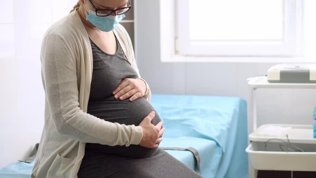 Young Pregnant Woman In Medicine Protective Mask On Hospital Couch Before Medical Control Cardiotocography. Visit Gynecologist Doctor At Clinic. Examine Expectant Belly Baby Mother Healthcare Check Up