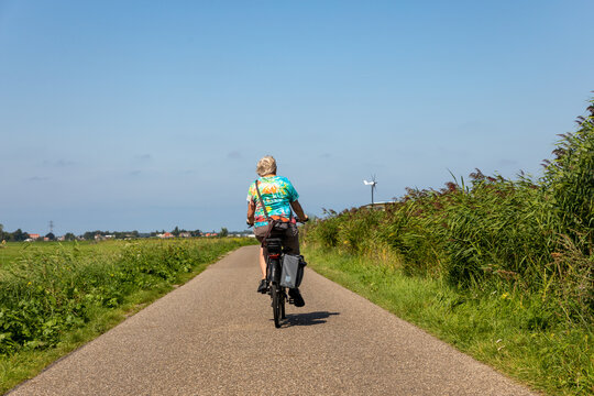 Elderly lady cycling on a asphalt road (dike) on a electric bike in the South-Holland village of Lisse in the Netherlands. Taken from the back. With a blue and sunny sky.