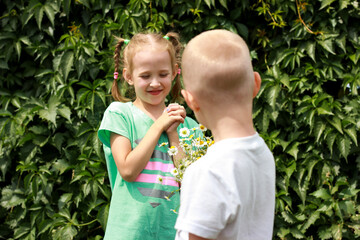 the boy stands with his back and gives a bouquet of daisies to the girl who opened her mouth with joy, blonde girl with pigtails from the back lifted up a bouquet, Valentines or mother day concept.