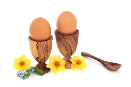 Healthy fresh brown eggs for breakfast in olive wood egg cups with narcissus and forget me not flowers. Health food concept for Easter and Spring, on white background.