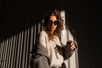 Sunlight portrait of a beautiful young girl with curly hair with stylish sunglasses in a fashionable coat and sweater walks on the street near a metal wall. Female casual style and fashion