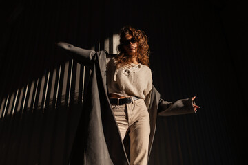 Stylish curly-haired girl with sunglasses in a fashionable long coat with a sweater and pants poses on the street near a metal wall in sunlight and shade