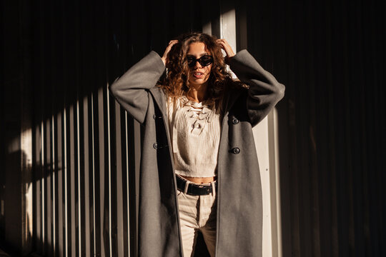 Stylish beautiful young curly-haired girl with fashion sunglasses in a fashionable long coat with a vintage sweater in the city in sunlight and shade
