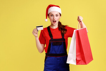 Excited astonished delivery woman with shopping bags and credit card in hands, paying with bank card, wearing blue overalls and santa claus hat. Indoor studio shot isolated on yellow background.