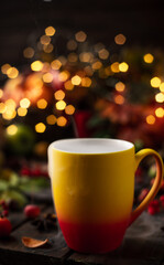 a red and yellow mug with a steaming drink. Fruits and spices are all around on a wooden table. Yellow lights of garlands are burning behind