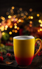a red and yellow mug with a steaming drink. Fruits and spices are all around on a wooden table. Yellow lights of garlands are burning behind