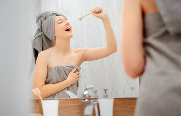 Fototapeta premium Young woman with a towel on her head brushes her teeth and sings as if the toothbrush is a microphone. Concept of dentistry and care.