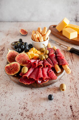 Prosciutto with figs, cheese, grapes, olives and breadsticks on a round board on a light gray background