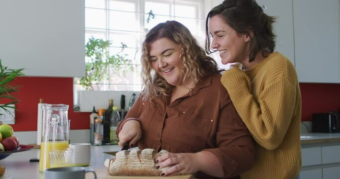 Happy caucasian lesbian couple slicing bread and laughing in kitchen
