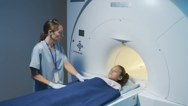 Slowmo shot of female MRI technician operating scanner while examining little girl in radiology room of modern clinic