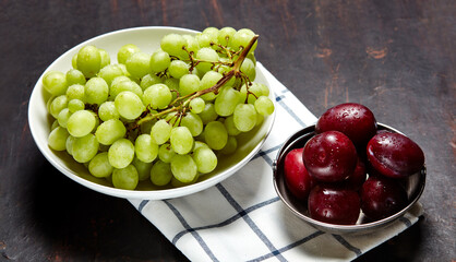 Branch of ripe green grape on plate with water drops and plums in bowl. Juicy fruits on wooden background, closeup. Healthy food on dark kitchen table