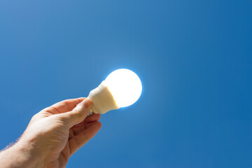 Hand holding a lit energy saving LED light bulb with a blue sky in the background.
