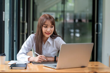 Fototapeta na wymiar Young Asian businesswoman working holding a pen using a taking note tablet looking at the laptop screen at the office.
