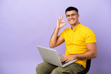 Young man sitting on a chair with laptop showing ok sign with fingers