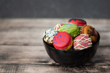 An enticing assortment of decorated macarons nestled in a dark bowl, contrasting with the rustic wood backdrop.