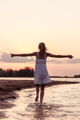 Fototapeta na wymiar Beautiful woman on the beach rear view. A young slender young blonde in a white dress runs along the river bank against the background of the sunset and with her hands raised. 