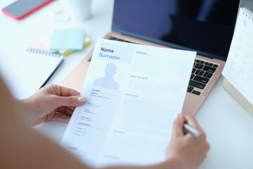 Woman sitting at table in front of laptop and holding paper with resume for employment in her hands