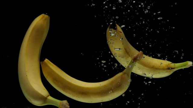Slow motion three bananas falling into transparent water on black background. Fresh fruits splashing in aquarium. Healthy food, diet, air bubbles