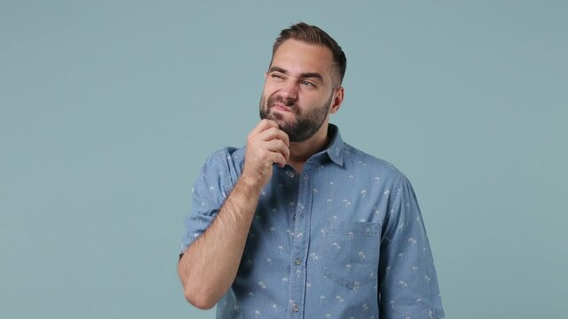 Fun insighted smart proactive pensive young brunet bearded man 20s wears denim shirt looks around thinks scratches at temple comes up with ideas raised finger isolated on pastel light blue background