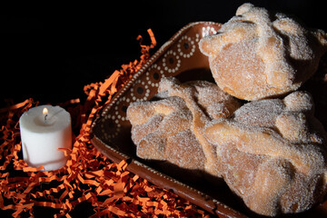 Tres piezas de pan de muerto sobre plato de barro con fondo naranja y negro y vela