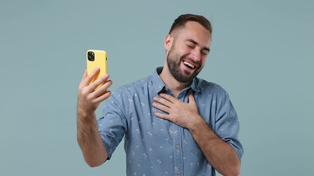 Excited young brunet bearded man 20s wear denim shirt get video call using mobile cell phone doing selfie talk conducting pleasant conversation greet with hand isolated on pastel light blue background