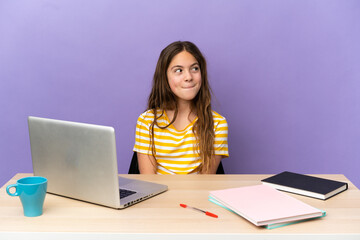 Little student girl in a workplace with a laptop isolated on purple background having doubts while looking up