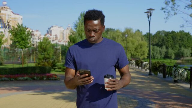 Thoughtful Afro-American man walks street looks around and navigates in smartphone holding cup with coffee or tea in hand. Pensive African adult male ethnic black wears blue t-shirt uses mobile phone