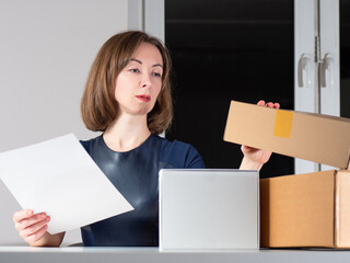 Woman next to cardboard boxes. She closes he box. Businesswoman holds a sheet of paper in her hand. Concept - she is engaged in home business. Woman home business owner. Girl on background of window