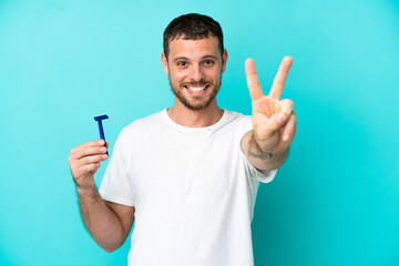 Brazilian man shaving his beard isolated on blue background smiling and showing victory sign