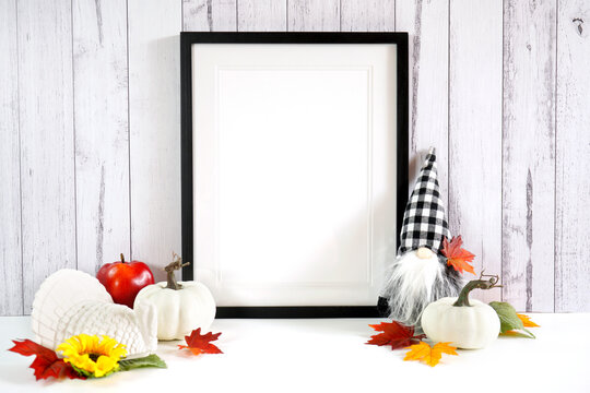 Artwork print poster frame product mockup. Thanksgiving farmhouse theme with gnome, turkey, white pumpkins and autumn fall leaves, on a white wood background. Negative copy space.
