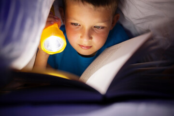 A young boy reading a book under the covers with a flashlight at dark night time - obrazy, fototapety, plakaty