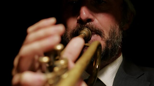Closeup of face of man in hat playing trumpet in spotlight, front view. Musician plays trumpet on dark background