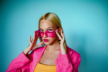 Fashion portrait of beautiful blonde woman wearing trendy pink sunglasses, fuchsia color blazer, orange top, big eaarings, posing on blue backgroumd. Copy, empty space for text
